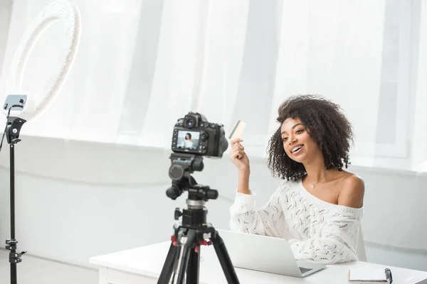 Selective focus of happy african american influencer in braces holding credit card near laptop and looking at digital camera — Stock Photo