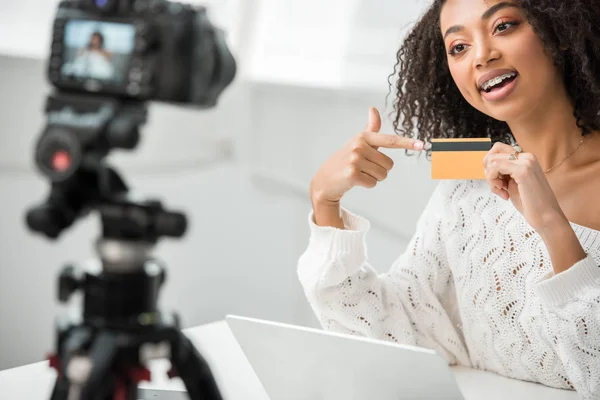 Selective focus of african american influencer in braces pointing with finger at credit card and looking at digital camera — Stock Photo