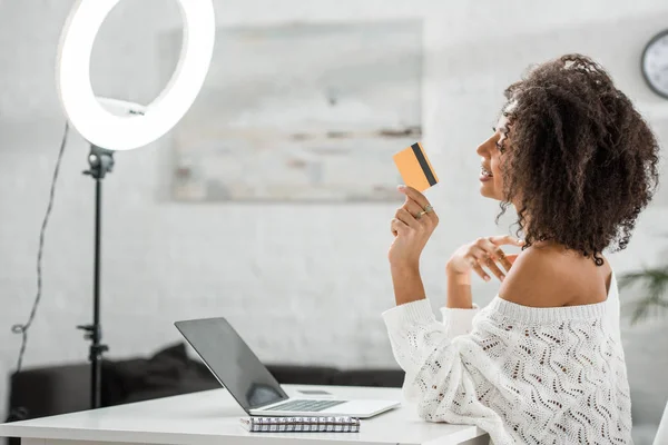 Vue latérale de heureux influenceur afro-américain dans les appareils tenant la carte de crédit près d'un ordinateur portable — Photo de stock