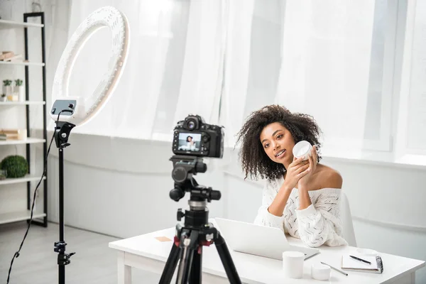 Selective focus of attractive african american influencer in braces holding container with cosmetic cream near digital camera — Stock Photo