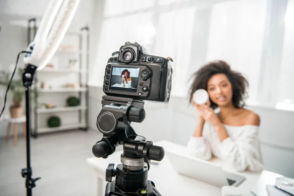 Selective focus of digital camera with attractive african american influencer in braces holding container with cosmetic cream on display — Stock Photo