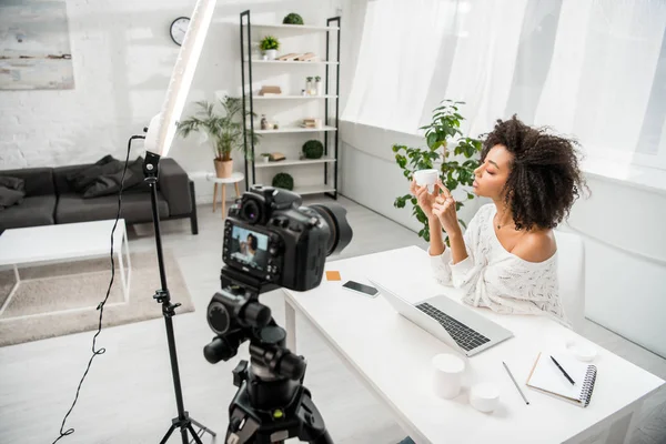 Foco seletivo do afro-americano influenciador segurando recipiente com creme cosmético perto da câmera digital — Fotografia de Stock