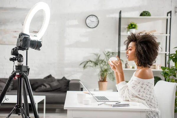 Vue latérale du jeune influenceur afro-américain tenant le récipient avec la crème cosmétique près de l'appareil photo numérique — Photo de stock