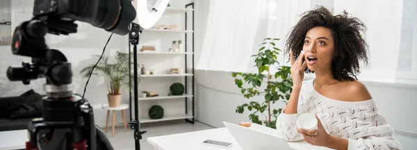 Panoramic shot of surprised african american influencer in braces applying cosmetic cream near digital camera — Stock Photo