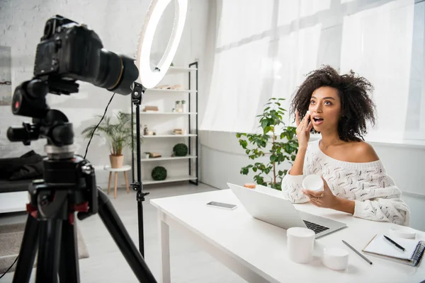 Selective focus of surprised african american influencer in braces applying cosmetic cream near digital camera — Stock Photo