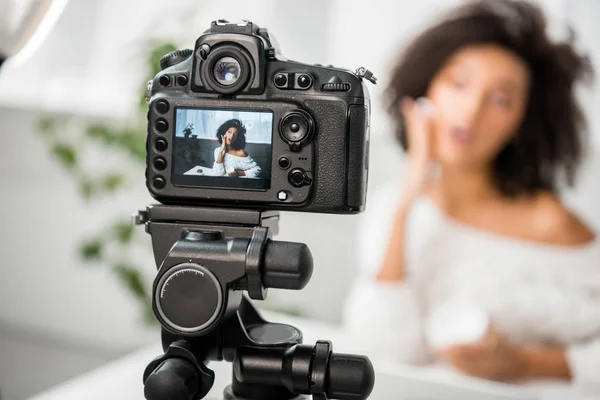 Enfoque selectivo de la cámara digital con el influencer afroamericano sorprendido aplicando crema facial en la pantalla - foto de stock