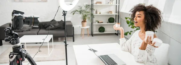 Panoramic shot of surprised african american influencer in braces holding containers with cosmetic cream near digital camera — Stock Photo