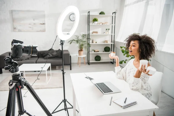 Surpreso afro-americano influenciador em aparelhos segurando recipientes com creme cosmético perto da câmera digital — Fotografia de Stock