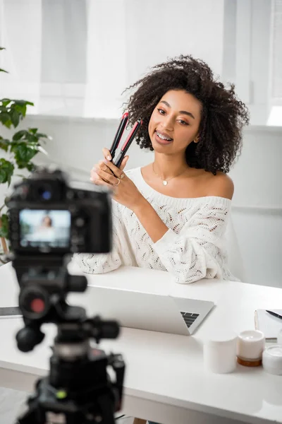Selective focus of happy african american influencer in braces holding hair straightener near laptop and digital camera — Stock Photo