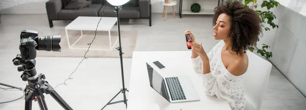 Panoramic shot of african american influencer holding hair straightener while talking near digital camera — Stock Photo