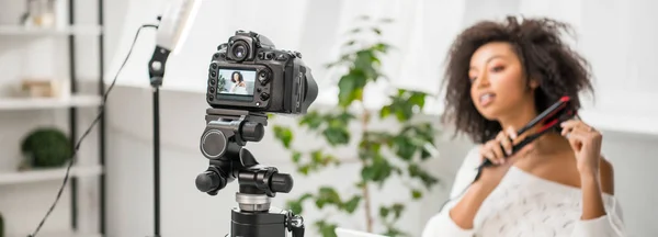 Panoramic shot of digital camera with african american influencer in braces using hair straightener on display — Stock Photo