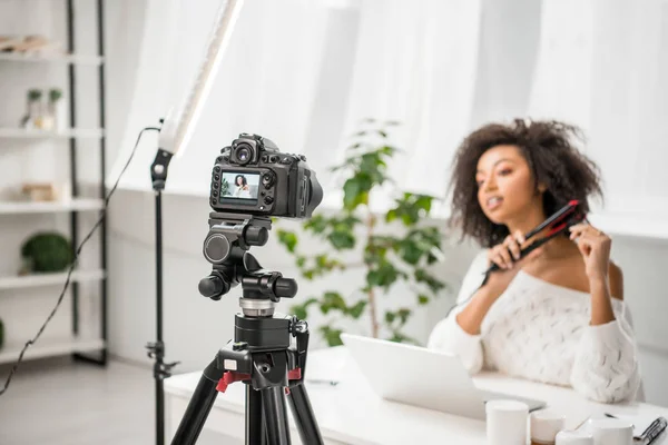 Selective focus of digital camera with african american influencer in braces using hair straightener on display — Stock Photo