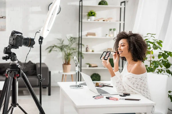 Influencer afroamericano feliz en aparatos ortopédicos que sostienen la paleta con sombra de ojos cerca de la cámara digital - foto de stock