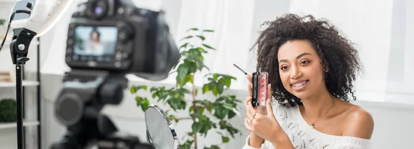Tiro panorâmico de feliz blogueiro de vídeo americano africano em aparelho segurando paleta com brilho labial perto da câmera digital — Fotografia de Stock