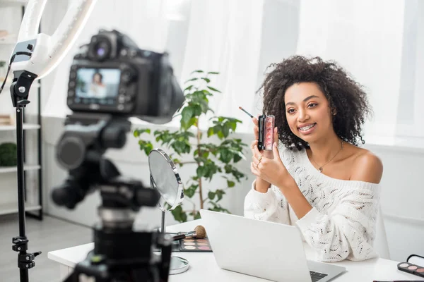 Foco seletivo de feliz blogueiro de vídeo americano africano em aparelho segurando paleta com brilho labial perto da câmera digital — Fotografia de Stock