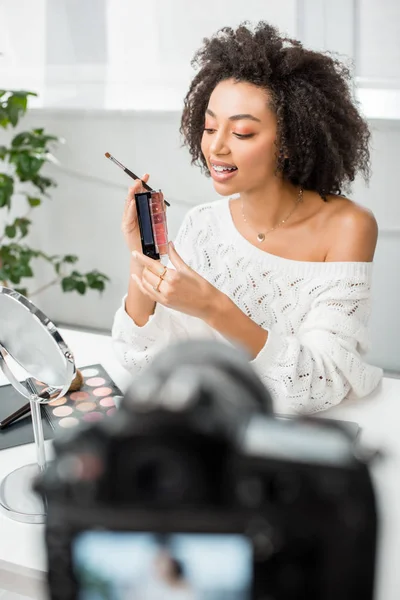 Foco seletivo de feliz blogueiro de vídeo americano africano em aparelho segurando paleta com brilho labial perto da câmera digital — Fotografia de Stock