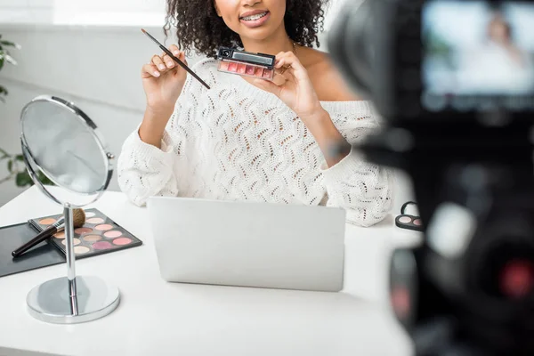 Vue recadrée du blogueur vidéo afro-américain heureux dans des bretelles tenant la palette avec du brillant à lèvres près de l'appareil photo numérique — Photo de stock