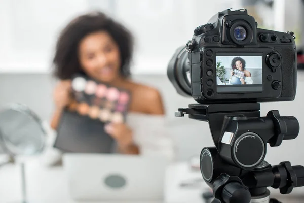 Mise au point sélective de l'appareil photo numérique avec heureuse fille afro-américaine dans des bretelles tenant palette avec ombre à paupières et pinceau cosmétique sur l'affichage — Photo de stock