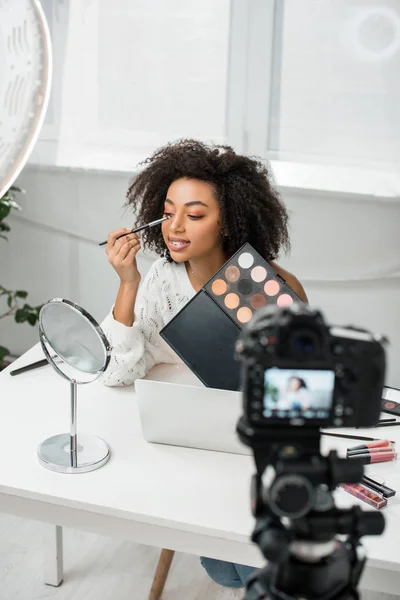 Foco seletivo de feliz blogueiro de vídeo americano africano em aparelhos aplicando sombra de olhos perto da câmera digital — Fotografia de Stock
