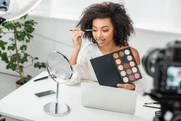 Selective focus of cheerful african american video blogger in braces applying eye shadow near digital camera — Stock Photo