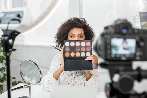 Enfoque selectivo de rizado afroamericano video blogger cubriendo la cara mientras sostiene la paleta de sombra de ojos cerca de la cámara digital - foto de stock