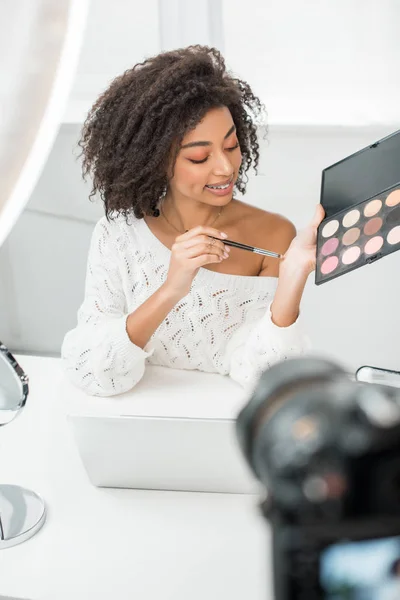 Foco selectivo del video blogger afroamericano feliz en aparatos ortopédicos que sostienen la paleta cosmética del cepillo y de la sombra de ojos cerca de cámara digital - foto de stock