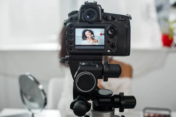 Selective focus of digital camera with happy african american video blogger in braces holding lip glosses and cosmetic brushes on display — Stock Photo