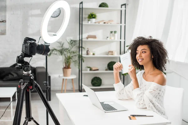 Heureux blogueur vidéo afro-américain dans les bretelles tenant carnet avec influenceur marketing lettrage près de l'appareil photo numérique — Photo de stock