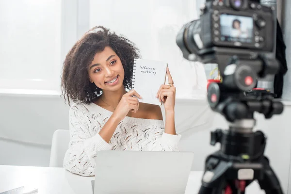 Foco seletivo de alegre blogueiro de vídeo americano africano em aparelho segurando notebook com letras de marketing influenciador perto da câmera digital — Fotografia de Stock