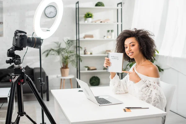 Feliz blogueiro de vídeo americano africano em aparelho apontando com o dedo para o notebook com letras de marketing influenciador perto da câmera digital — Fotografia de Stock