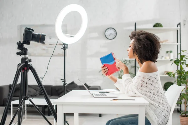 Side view of attractive african american influencer holding colorful box and talking near digital camera — Stock Photo