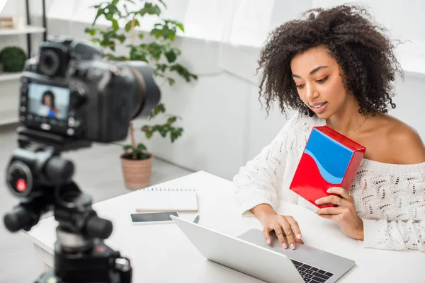 Selective focus of attractive african american influencer in braces holding colorful box and using laptop near digital camera — Stock Photo