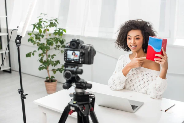 Selective focus of happy african american influencer in braces pointing with finger at colorful box near digital camera — Stock Photo