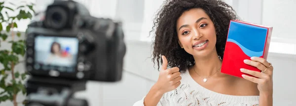 Panoramic shot of happy african american influencer in braces holding colorful box and showing thumb up near digital camera — Stock Photo