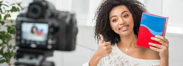 Panoramaaufnahme eines glücklichen afrikanisch-amerikanischen Influencers in Hosenträgern, der eine bunte Schachtel hält und den Daumen in der Nähe der Digitalkamera zeigt — Stockfoto