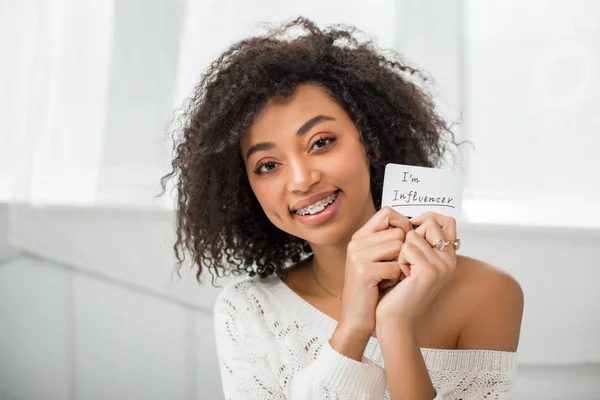 Gai afro-américaine fille dans bretelles tenant la carte avec je suis influenceur lettrage à la maison — Photo de stock
