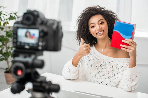 Messa a fuoco selettiva di felice influencer afro-americano in bretelle con scatola colorata e mostrando pollice verso l'alto vicino alla fotocamera digitale — Foto stock