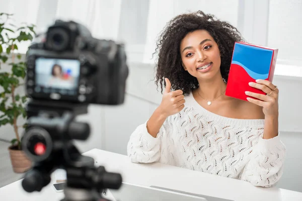 Selektiver Fokus glücklicher afrikanisch-amerikanischer Influencer in Hosenträgern, die bunte Schachtel in der Hand halten und Daumen hoch in der Nähe der Digitalkamera zeigen — Stockfoto