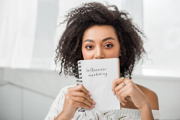 Encaracolado Africano americano menina segurando notebook com influenciador marketing lettering em casa — Fotografia de Stock