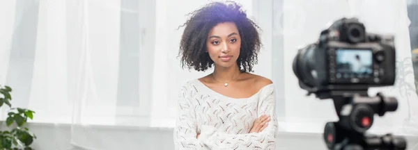 Panoramic shot of curly african american influencer standing with crossed arms and looking at digital camera — Stock Photo