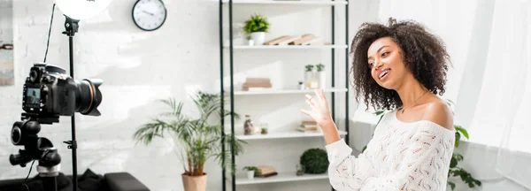 Panoramic shot of happy african american influencer in braces waving hand and talking near digital camera — Stock Photo