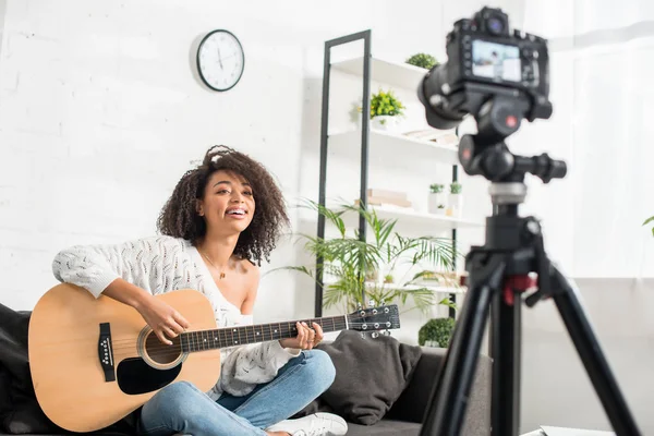 Foco seletivo da menina americana africana feliz em aparelho tocando guitarra acústica perto da câmera digital — Fotografia de Stock