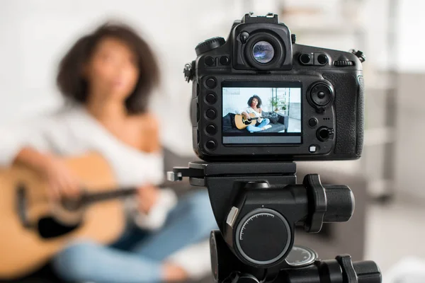Messa a fuoco selettiva della fotocamera digitale con giovane e riccia ragazza africana americana che suona la chitarra acustica in mostra — Foto stock