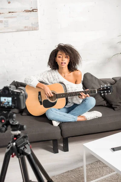 Enfoque selectivo de la joven afroamericana cantando y tocando la guitarra acústica cerca de la cámara digital - foto de stock