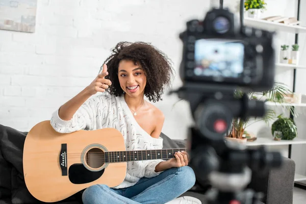 Messa a fuoco selettiva di allegra ragazza afroamericana in bretelle con chitarra acustica e puntando con il dito alla fotocamera digitale — Foto stock