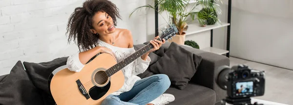 Панорамный снимок веселой африканской девушки в брекетах, держащей акустическую гитару и смотрящей на цифровую камеру — стоковое фото