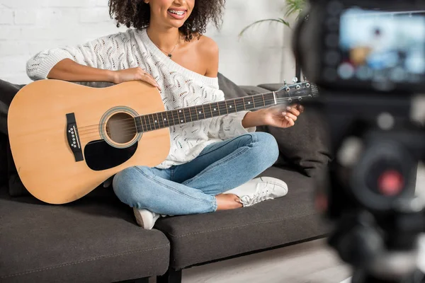 Abgeschnittene Ansicht eines fröhlichen afrikanisch-amerikanischen Mädchens in Hosenträgern, das eine Akustikgitarre in der Nähe der Digitalkamera hält — Stockfoto