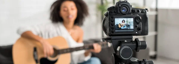 Panoramic shot of digital camera with young african american girl in braces playing acoustic guitar and singing on display — Stock Photo