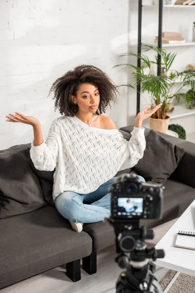 Enfoque selectivo del joven afroamericano influencer sentado en el sofá y haciendo gestos cerca de la cámara digital - foto de stock