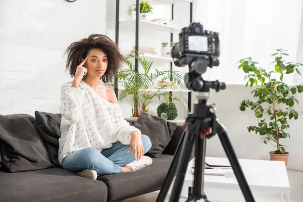 Selective focus of young african american influencer sitting on sofa and pointing with finger at head near digital camera — Stock Photo